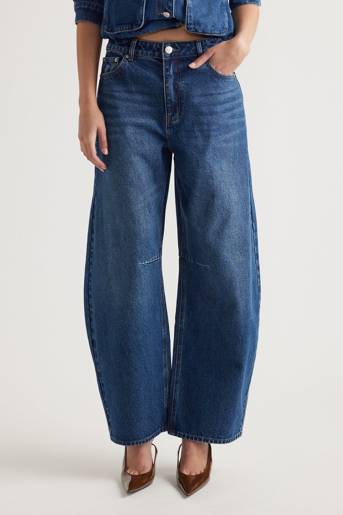 Woman wearing mid-rise wide-leg indigo jeans paired with pointed brown heels against a plain background.