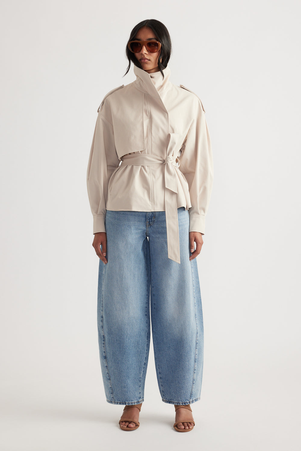 Front view of woman wearing light blue wide-leg mid rise jeans paired with beige belted jacket and brown sandals