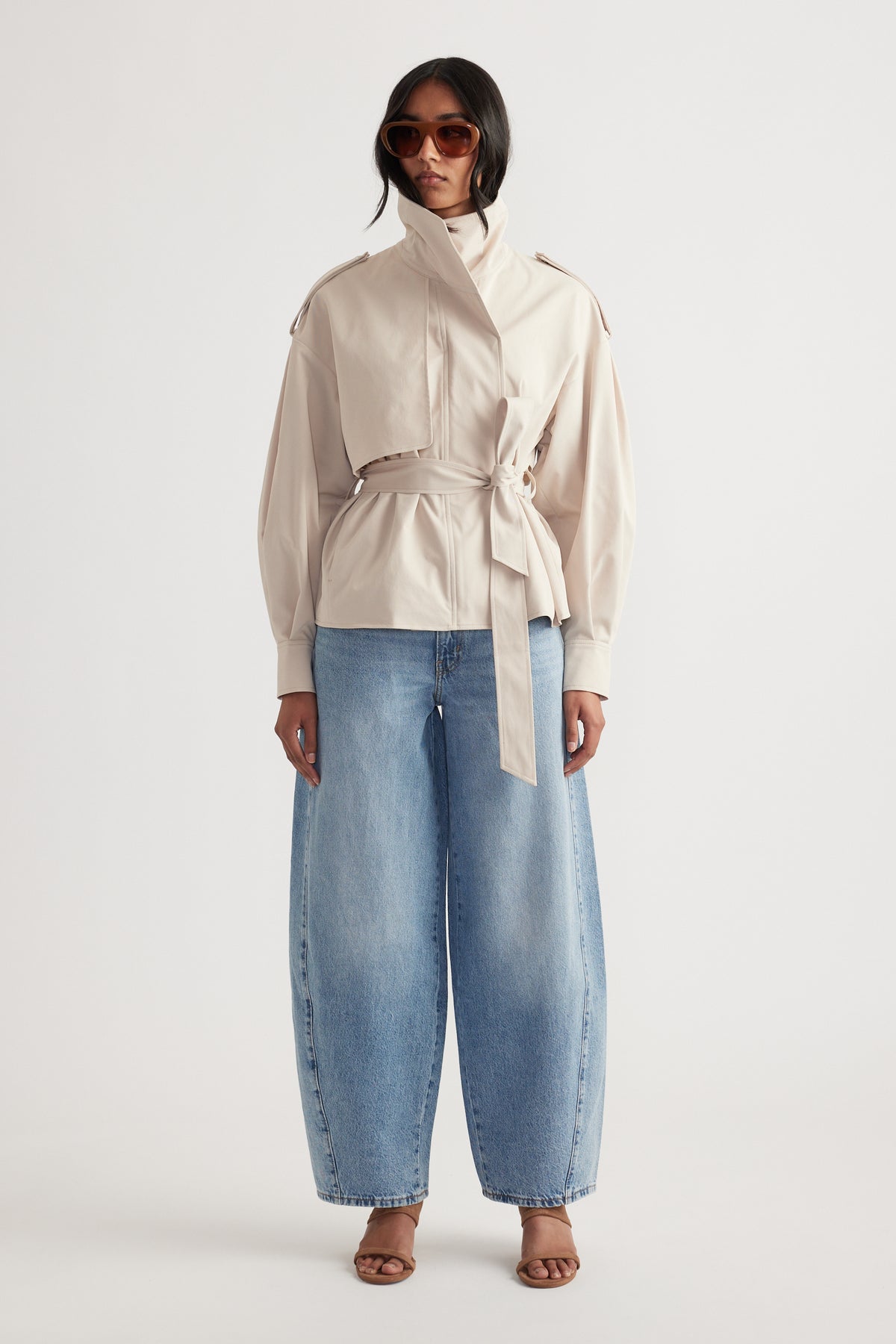 Front view of woman wearing light blue wide-leg mid rise jeans paired with beige belted jacket and brown sandals