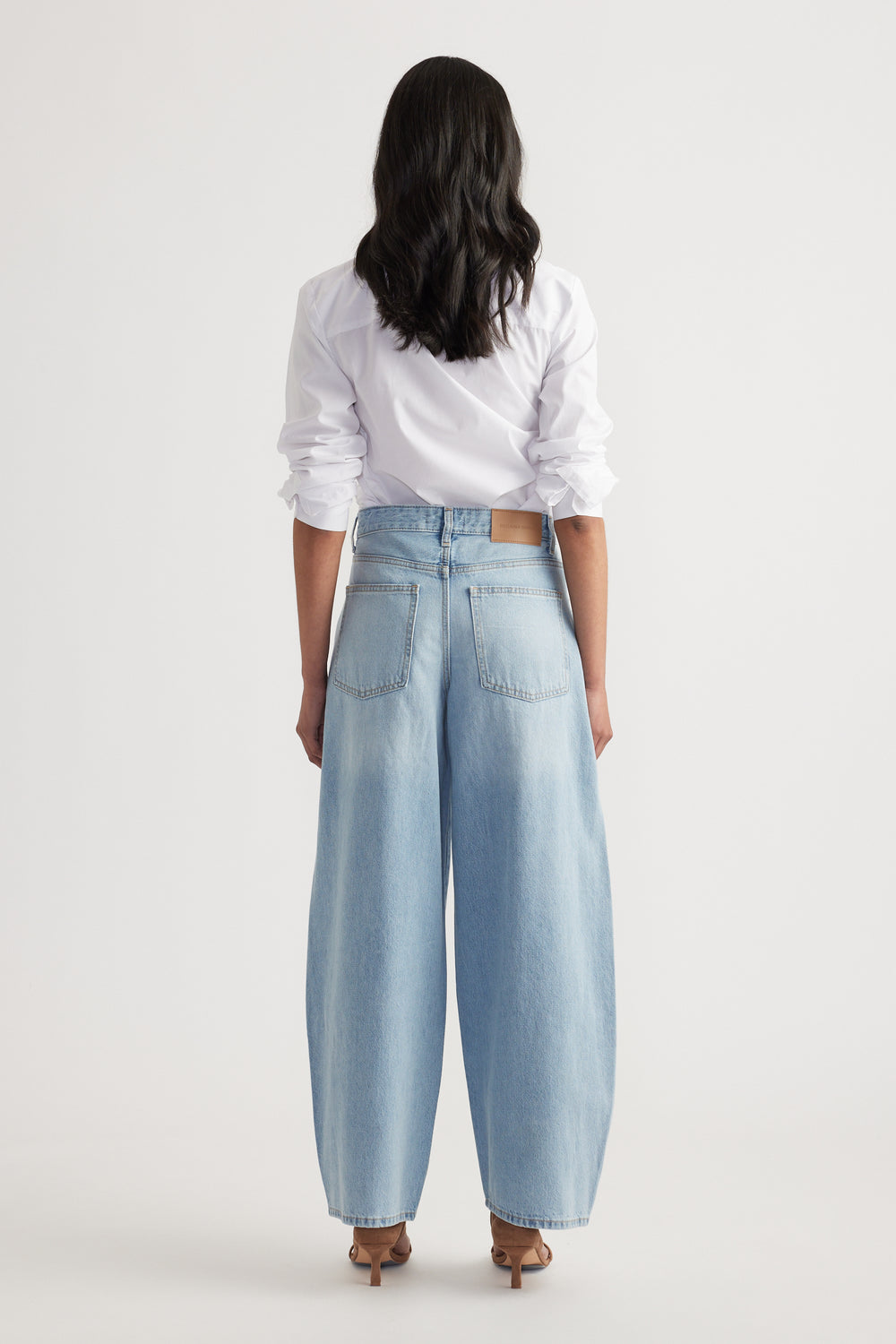 Woman wearing light blue high-waisted barrel-leg jeans paired with a white long-sleeve shirt and heels, back view