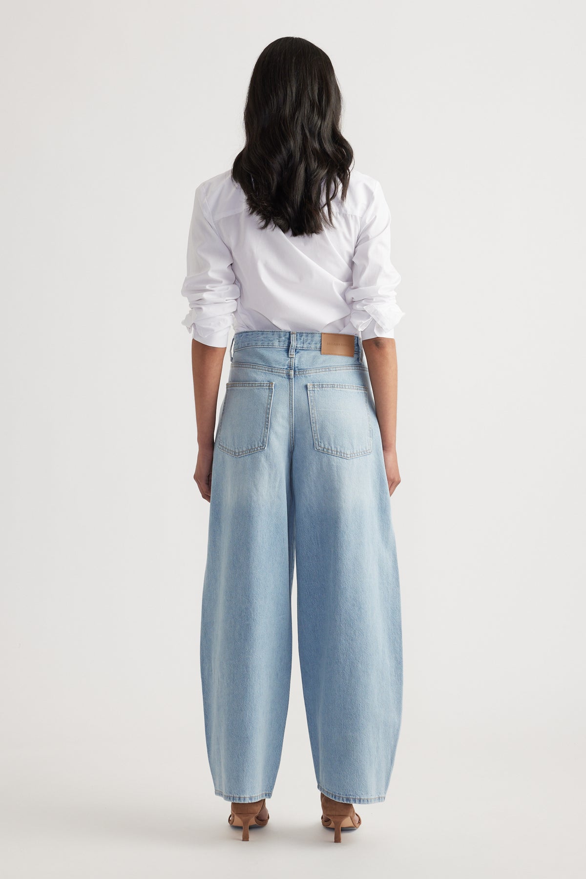 Woman wearing light blue high-waisted barrel-leg jeans paired with a white long-sleeve shirt and heels, back view