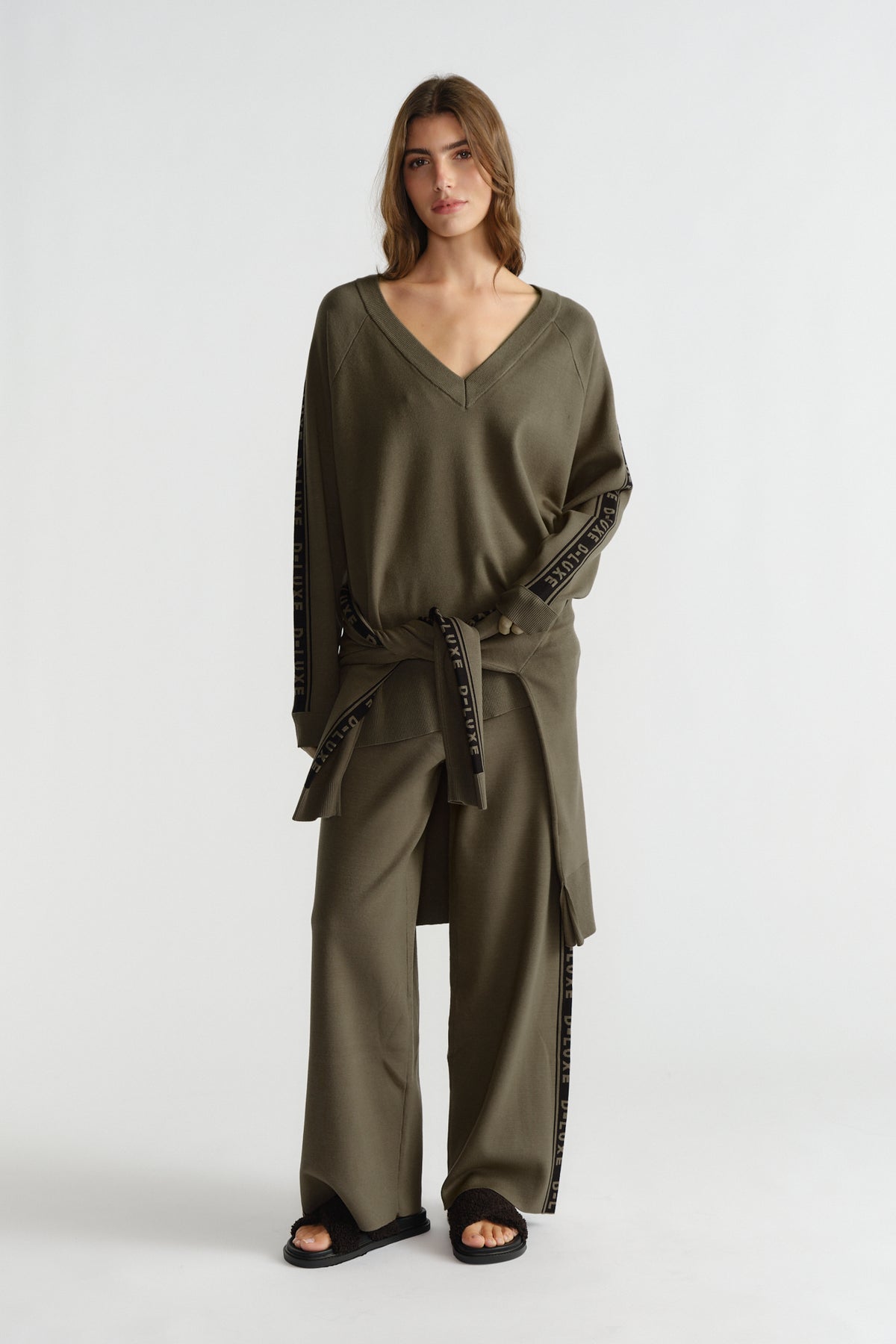 Woman wearing relaxed olive green knit top with matching wide-leg pants featuring side logo panels and tie detail.
