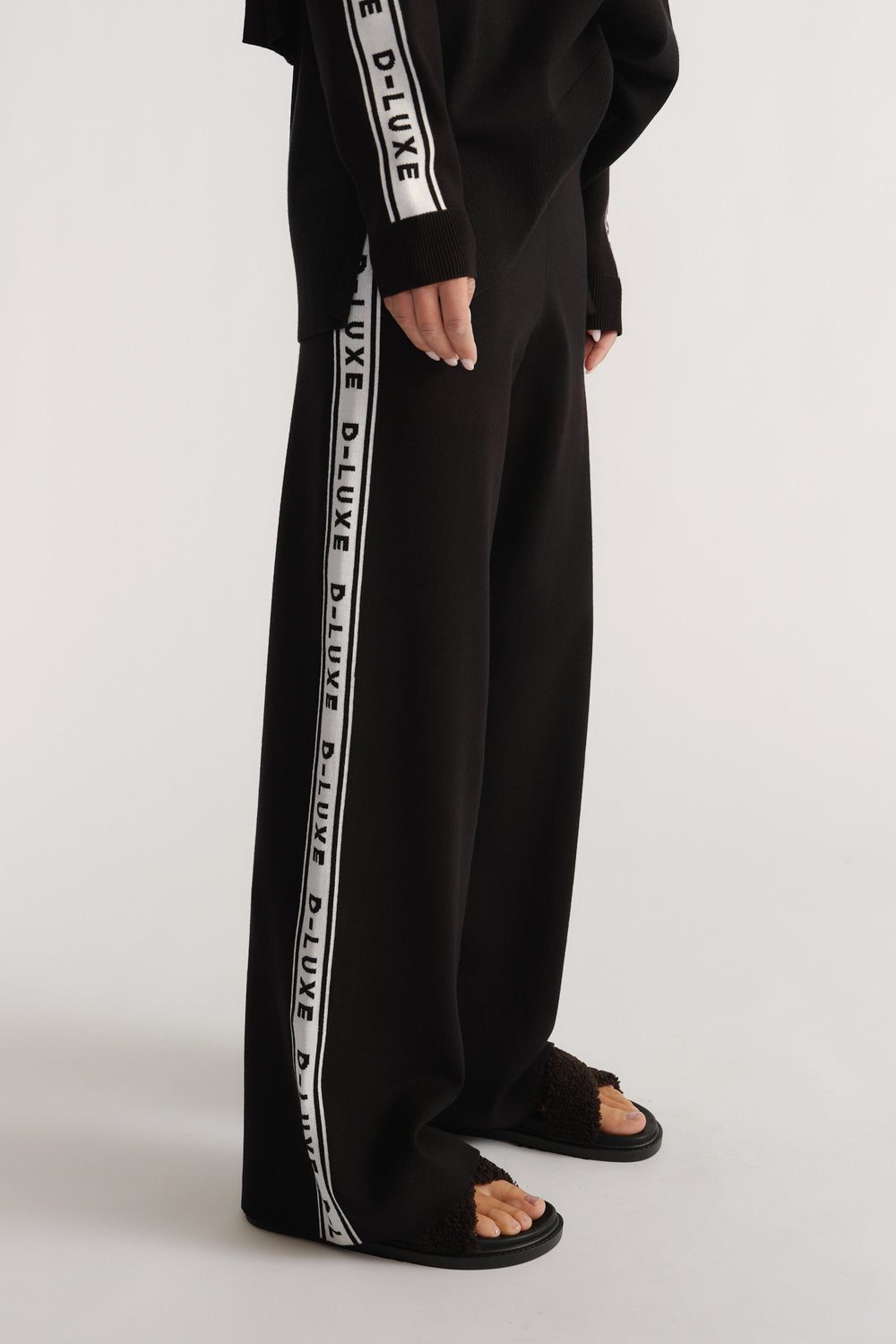 Side view of black knit pants with white logo panels, styled with matching long-sleeve top and textured sandals
