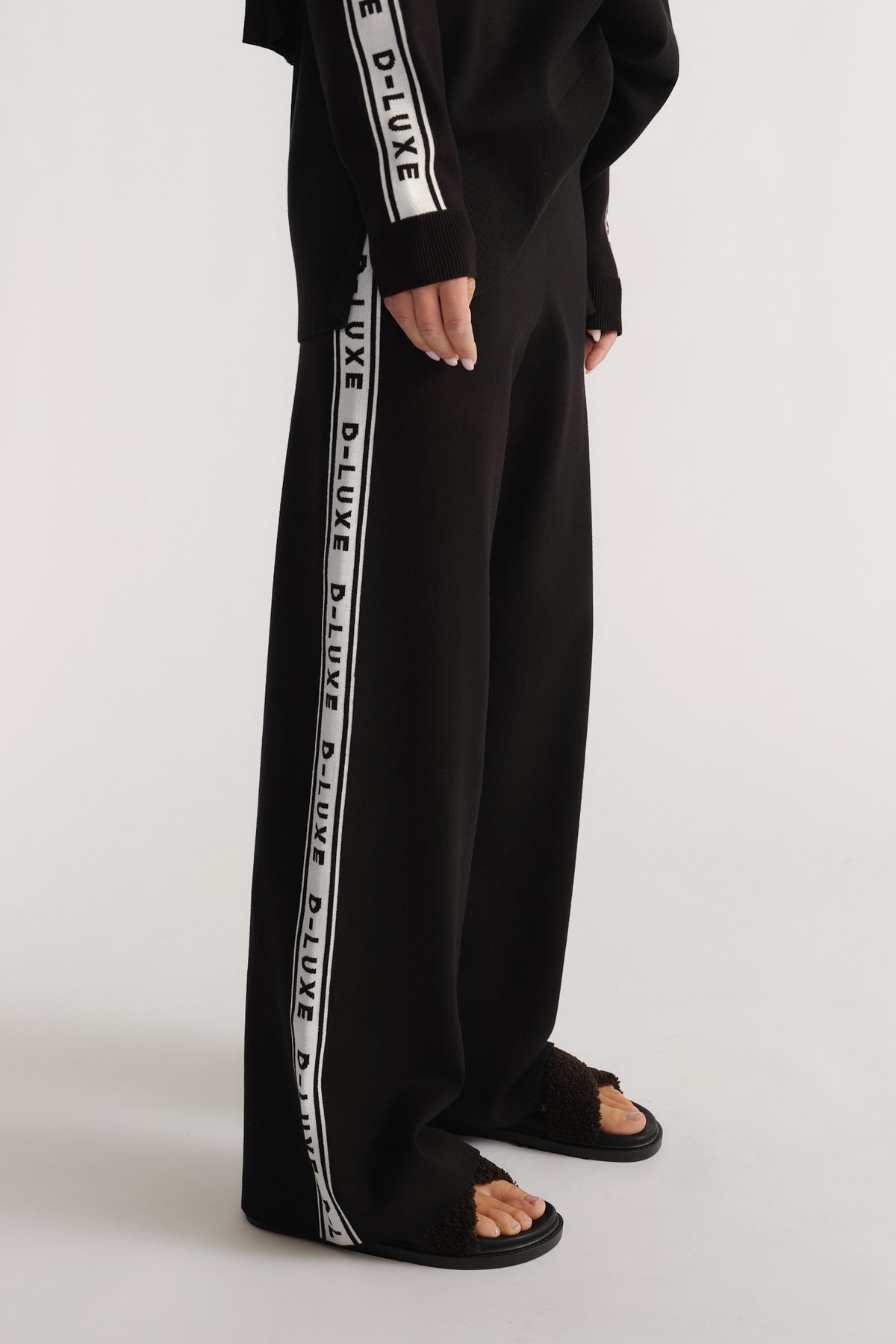 Side view of black knit pants with white logo panels, styled with matching long-sleeve top and textured sandals