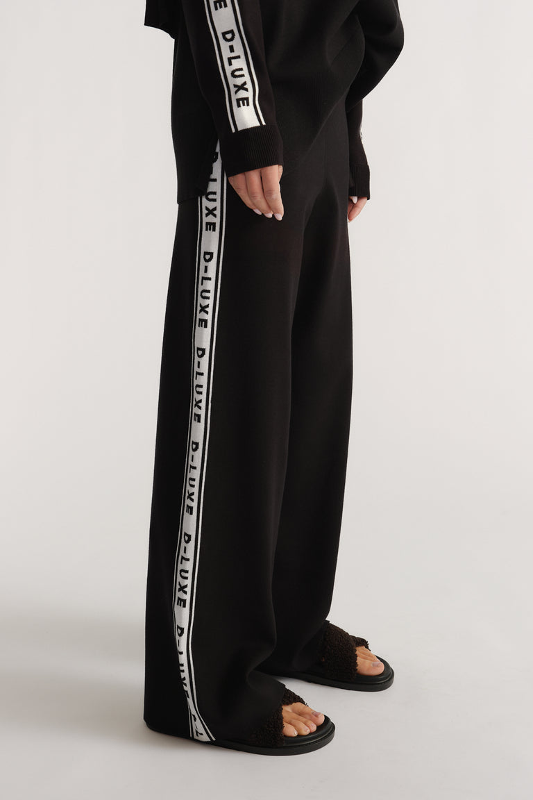 Side view of black knit pants with white logo panels, styled with matching long-sleeve top and textured sandals