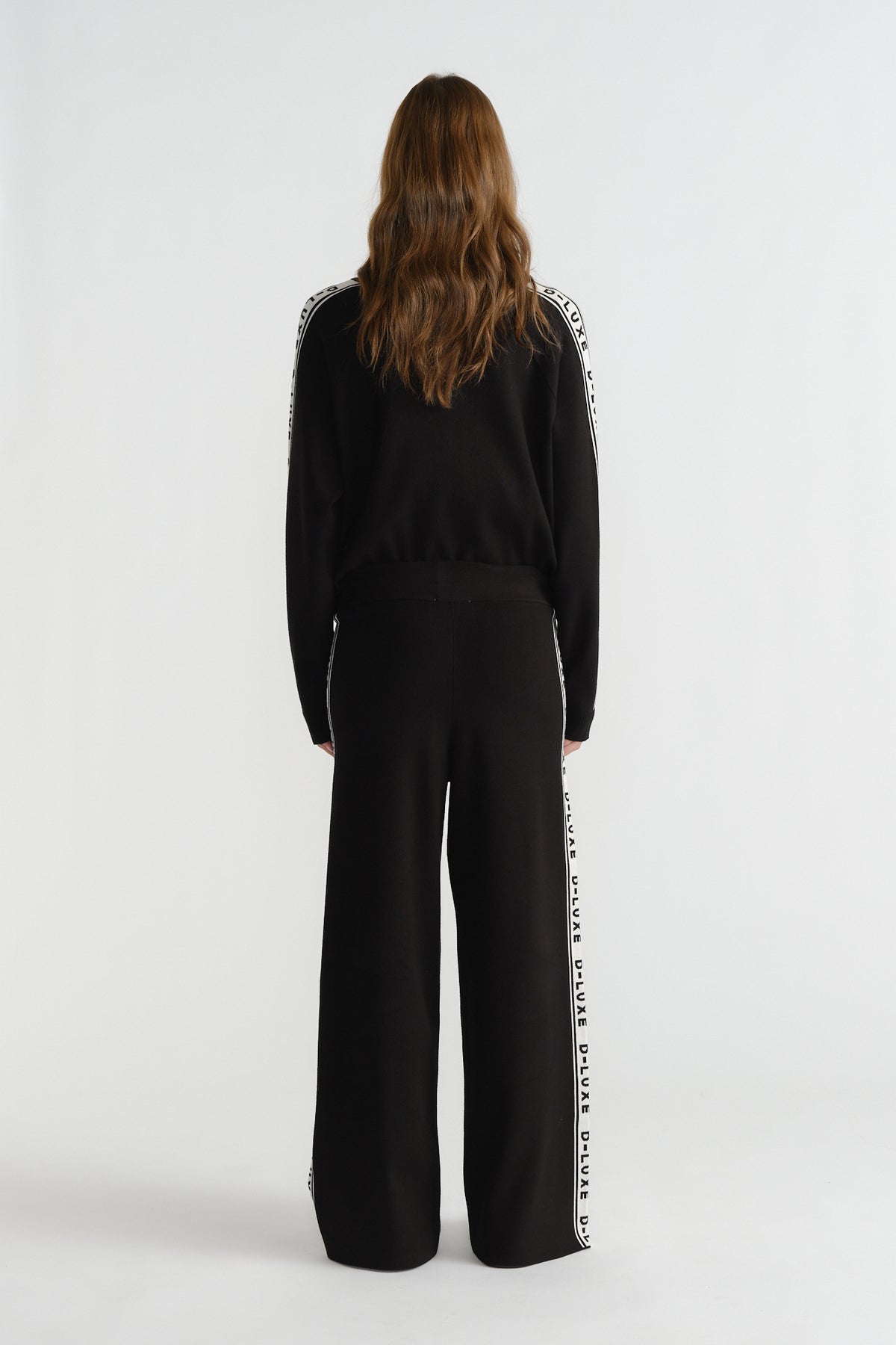 Back view of woman wearing black knit pants with side logo panel paired with matching long-sleeve top