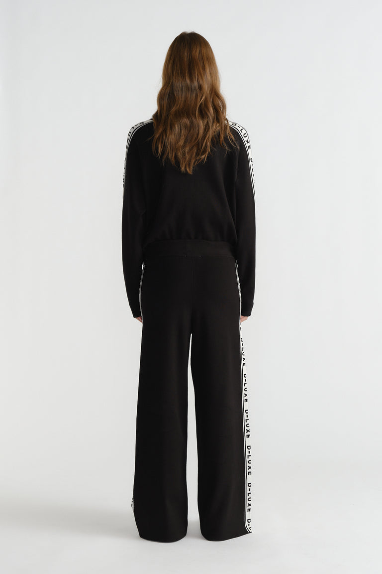 Back view of woman wearing black knit pants with side logo panel paired with matching long-sleeve top