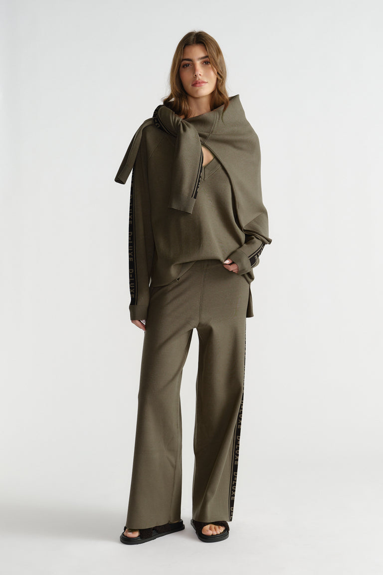 Woman wearing relaxed olive knit pants with side logo panels and matching knit top with tied sweater over shoulders