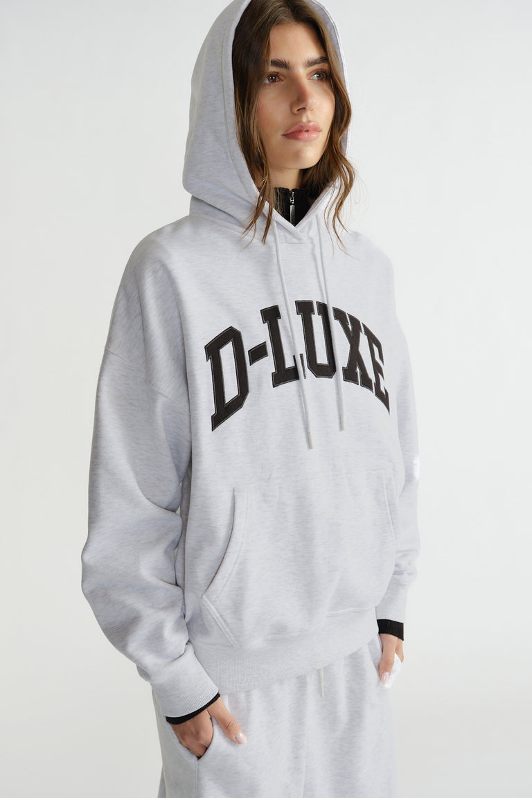 Woman wearing a light grey hoodie with bold lettering, featuring a relaxed fit and kangaroo pocket, styled casually.