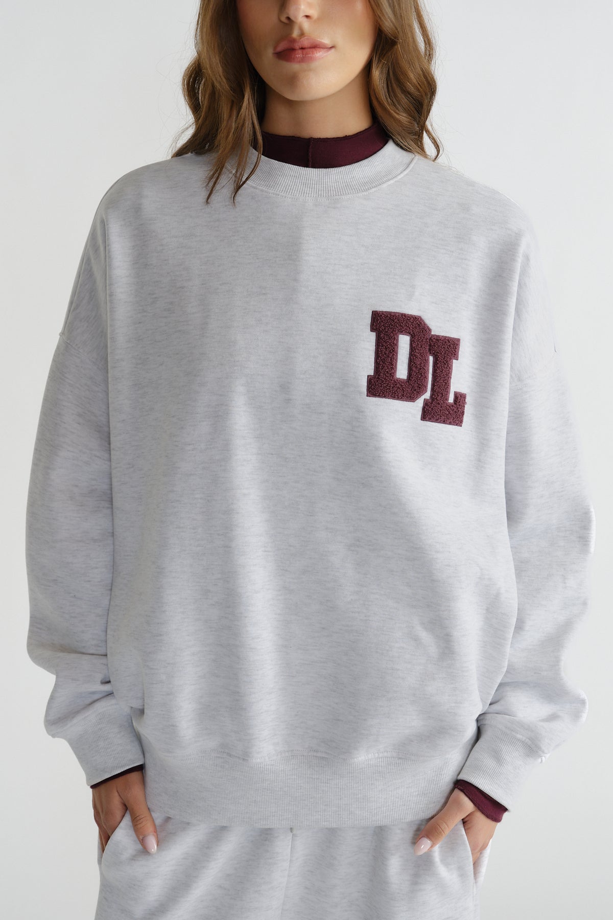 Light grey crew sweatshirt with relaxed fit, featuring textured maroon "DL" logo patch on the chest.