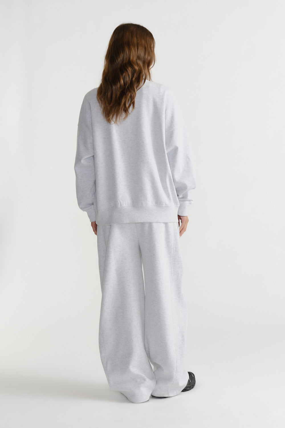 Model seen from behind wearing a relaxed light grey crewneck sweatshirt and matching wide-leg sweatpants.