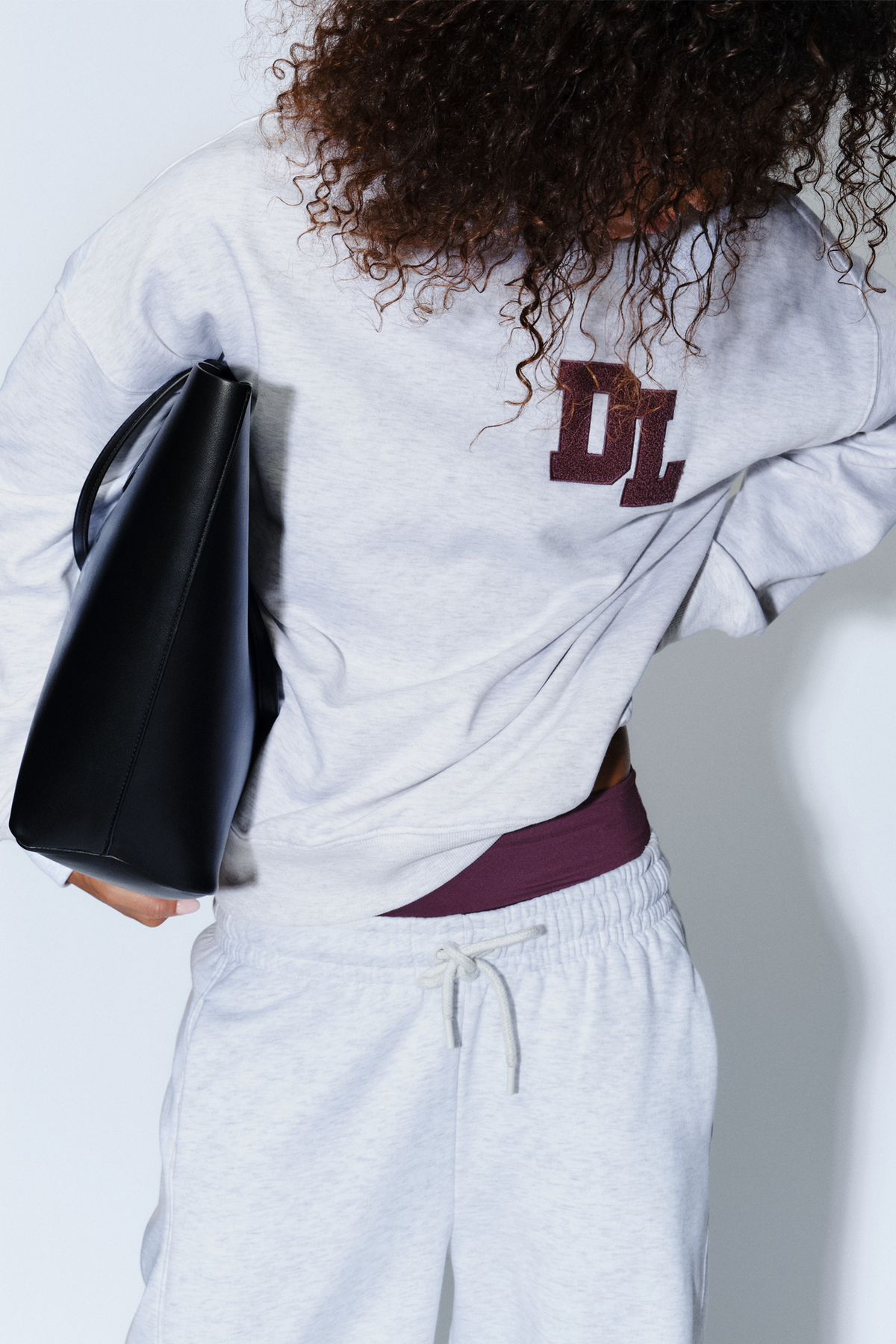 Person wearing a light grey crew sweat with a subtle logo, paired with matching soft lounge pants and a black handbag