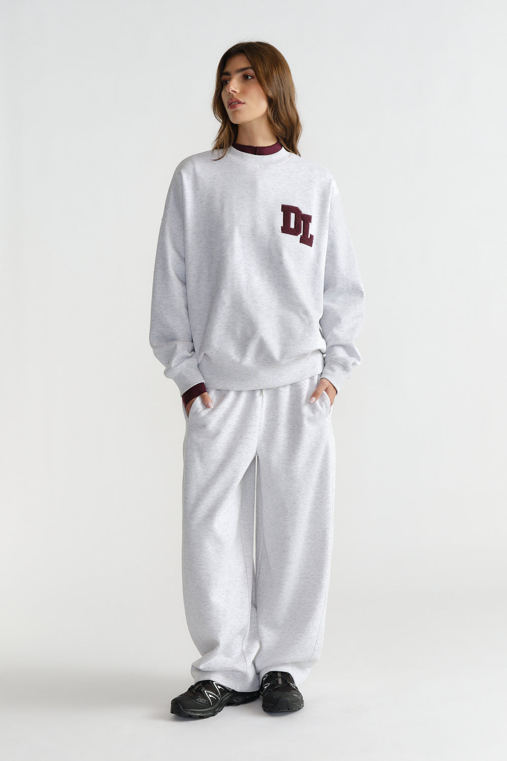 Woman wearing relaxed light grey barrel pants and matching sweatshirt with maroon logo detail and black sneakers