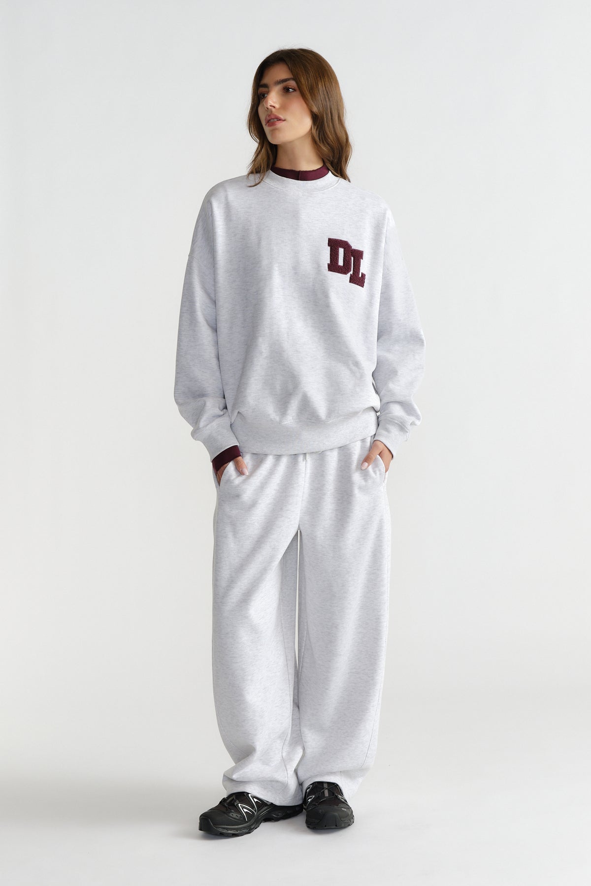 Woman wearing relaxed light grey barrel pants and matching sweatshirt with maroon logo detail and black sneakers