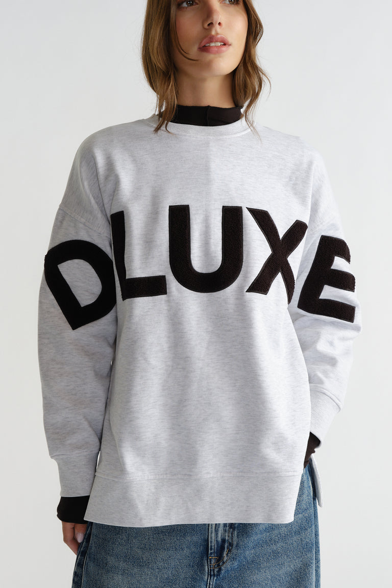 Woman wearing a light grey sweatshirt with bold black textured lettering across the chest, styled with casual denim jeans