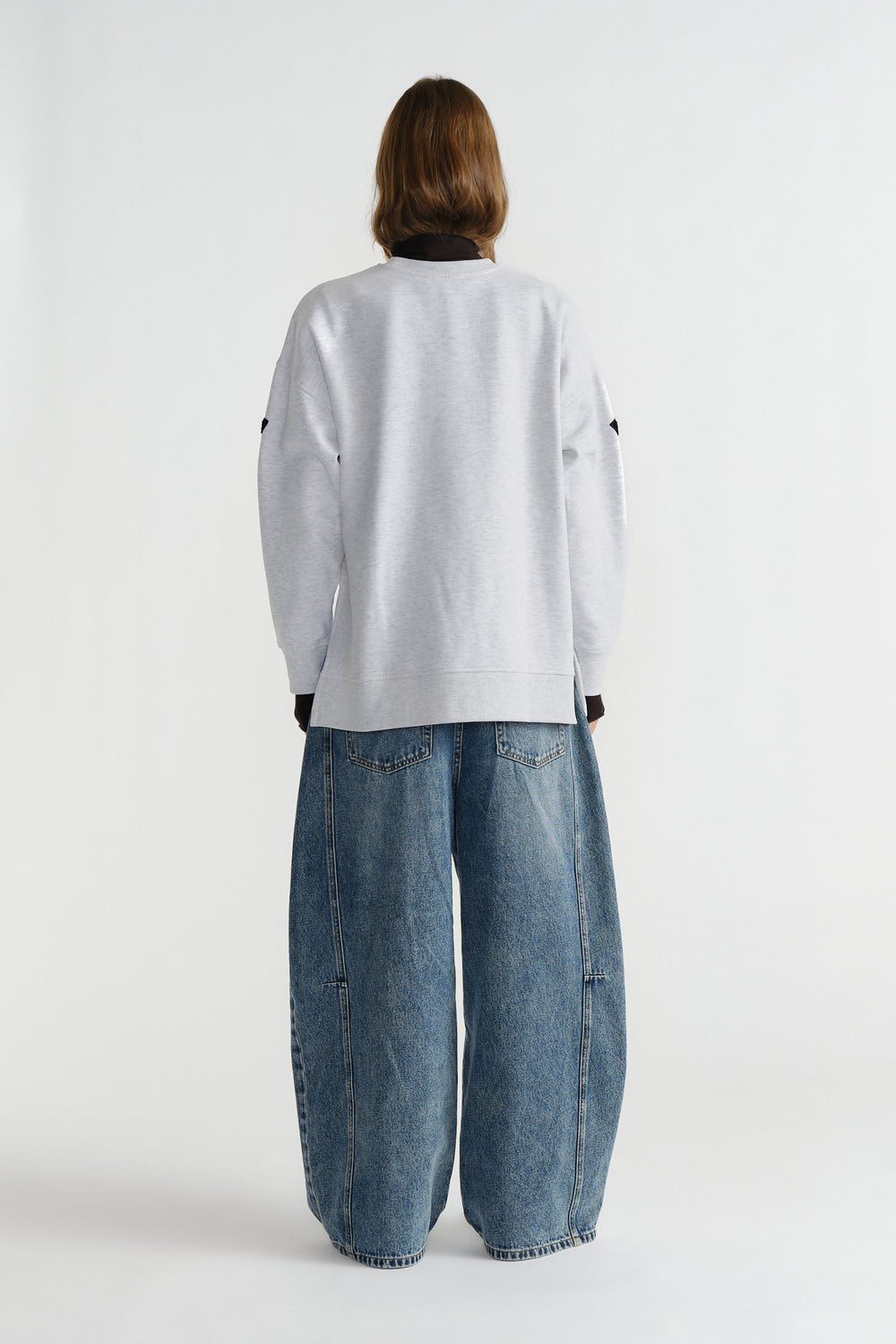 Back view of a model wearing a light grey relaxed sweat paired with wide-leg blue denim jeans against a white background