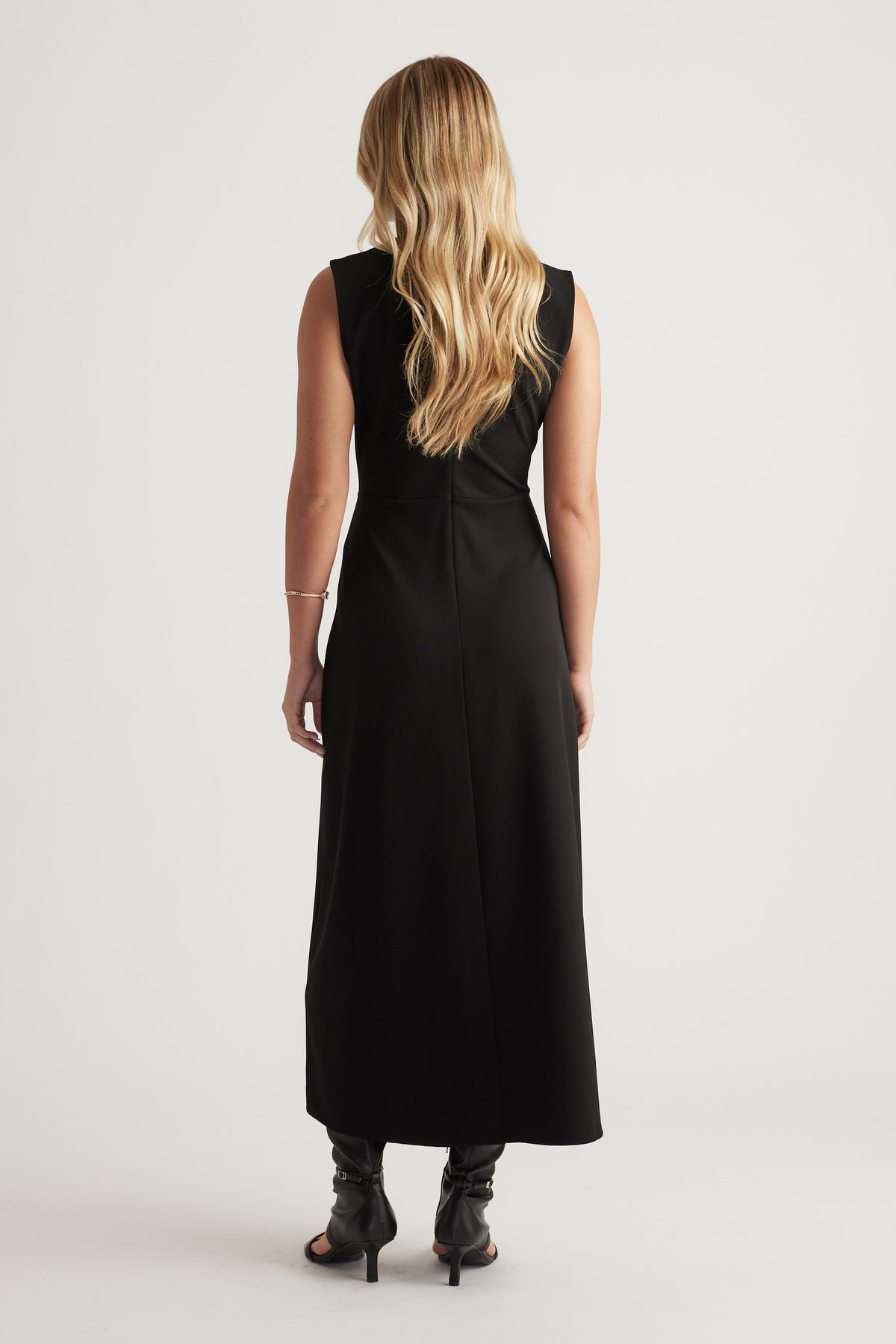 Back view of a woman wearing a sleeveless black midi dress paired with black heeled boots and loose blonde hair