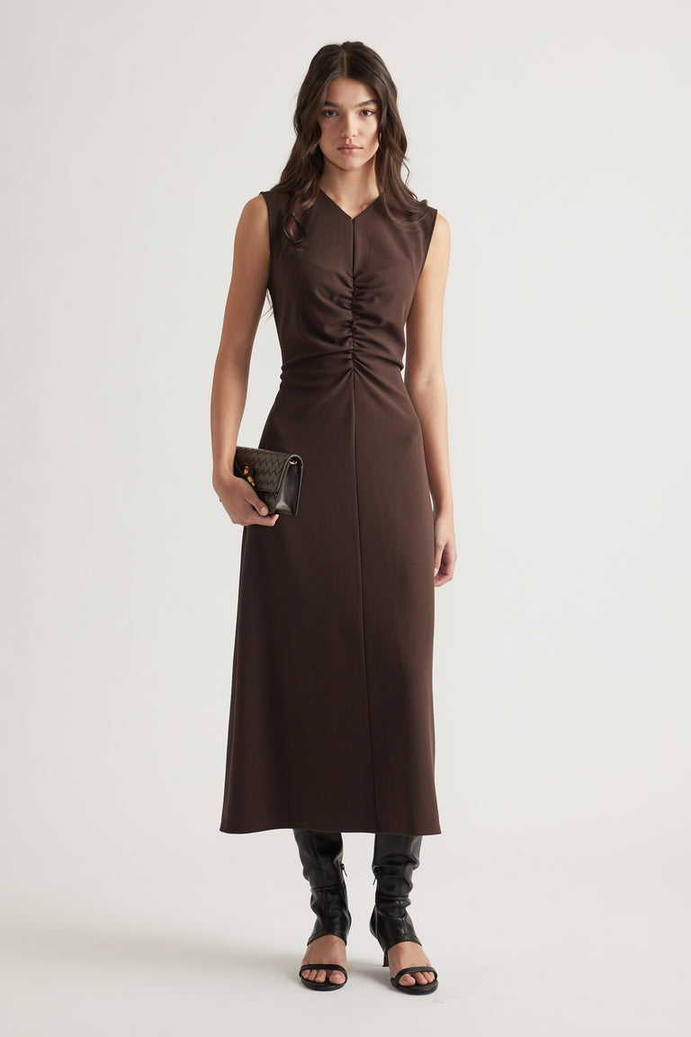 Woman wearing a sleeveless chocolate brown ruched midi dress styled with black heeled sandals and a quilted clutch bag