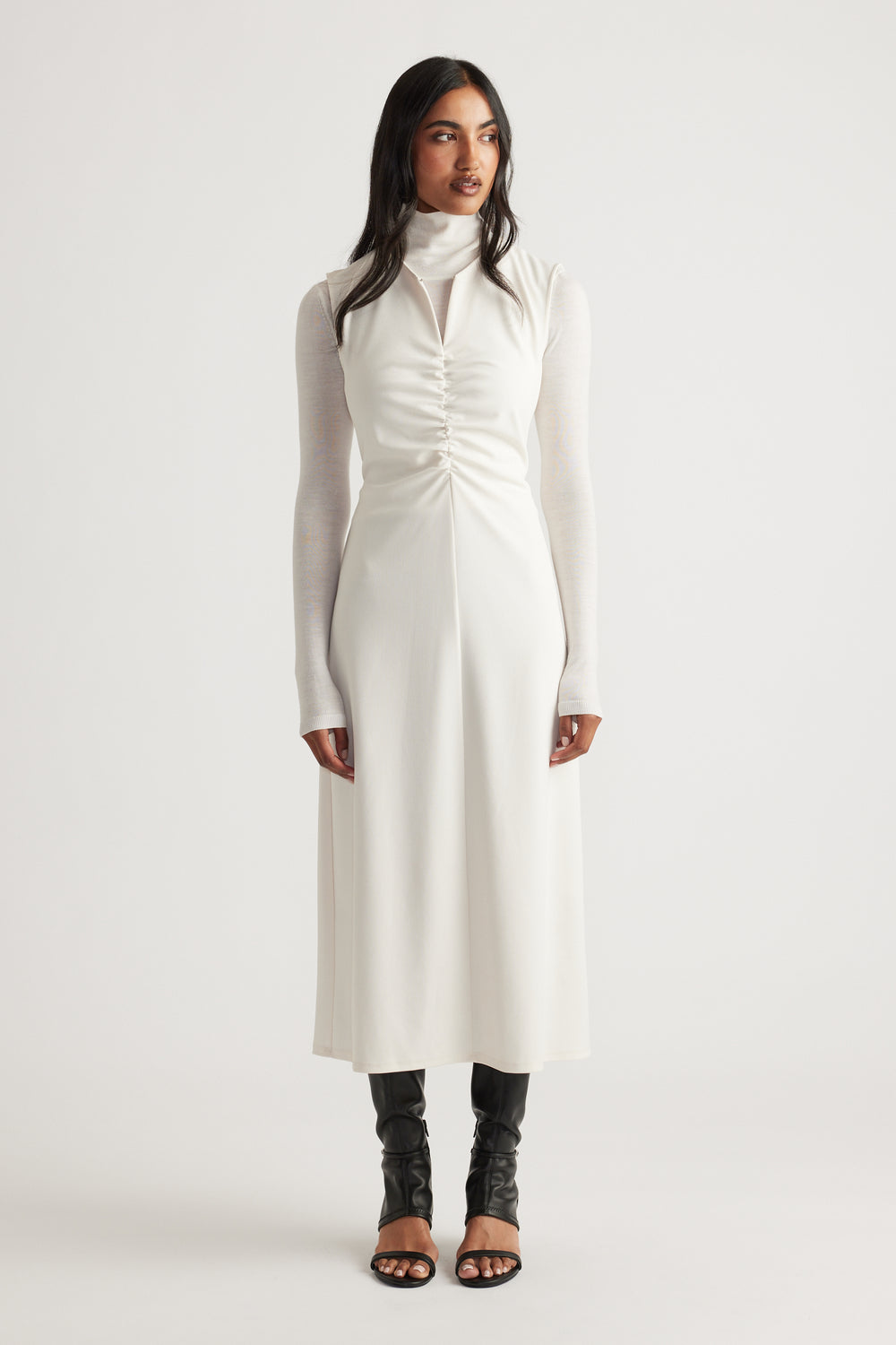 Woman wearing a white ruched midi dress layered over a fitted long-sleeve turtleneck and paired with black open-toe boots