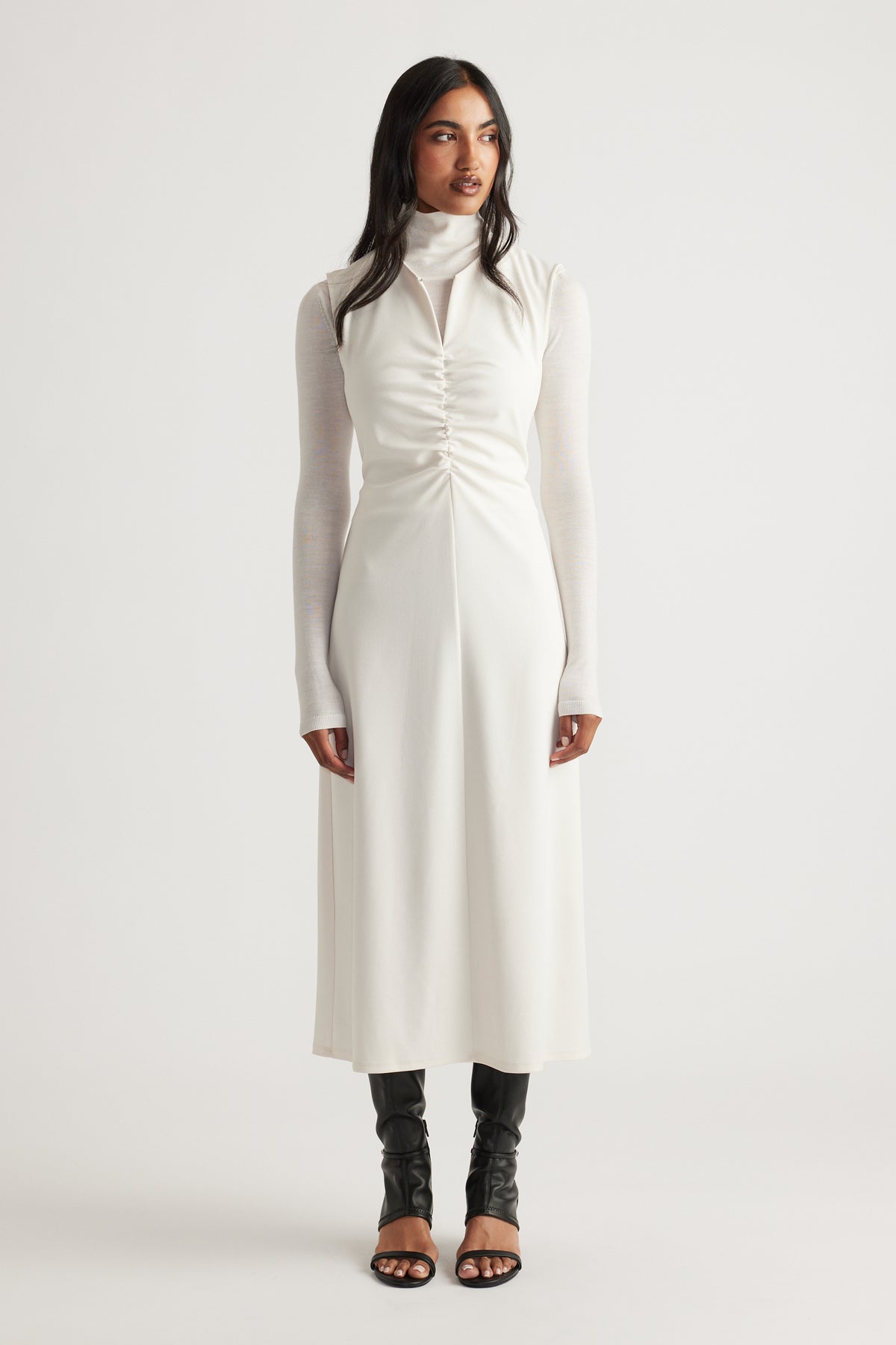 Woman wearing a white ruched midi dress layered over a fitted long-sleeve turtleneck and paired with black open-toe boots