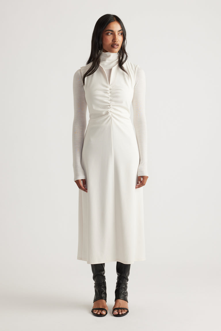 Woman wearing a white ruched midi dress layered over a fitted long-sleeve turtleneck and paired with black open-toe boots