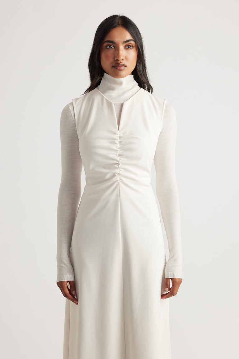 Woman wearing a white midi dress with ruched front detail layered over a fitted long sleeve turtleneck top