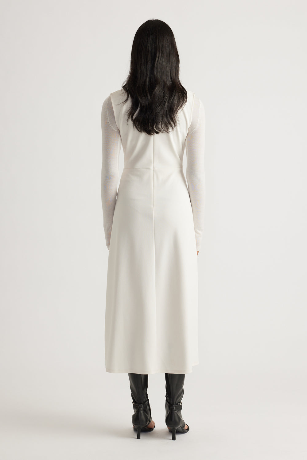 Back view of a sleeveless white midi dress worn over a sheer long-sleeve top paired with black heeled boots