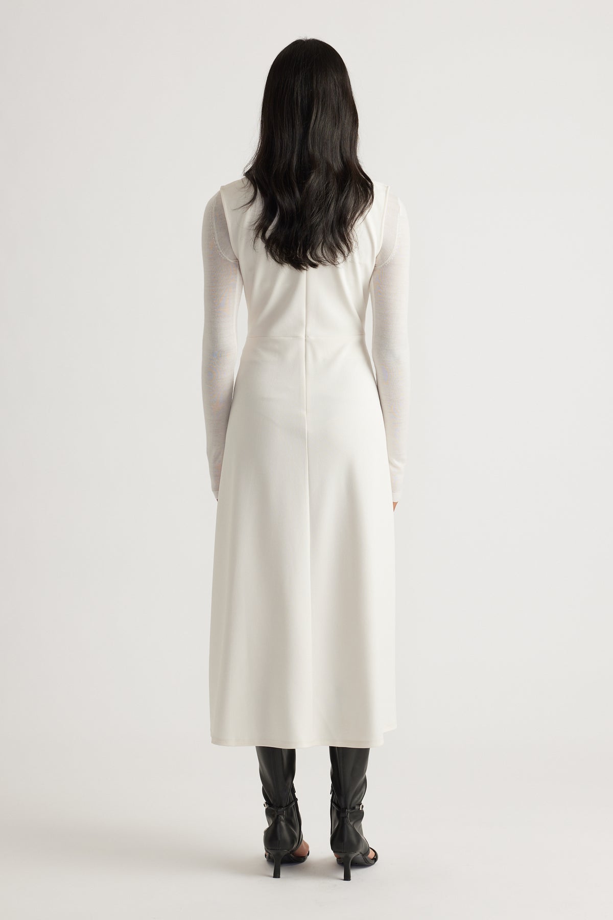 Back view of a sleeveless white midi dress worn over a sheer long-sleeve top paired with black heeled boots