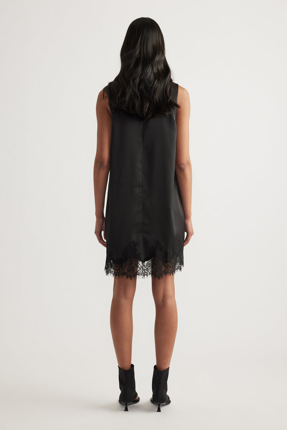 Woman viewed from behind wearing a black sleeveless shift mini dress with lace trim and ankle boots