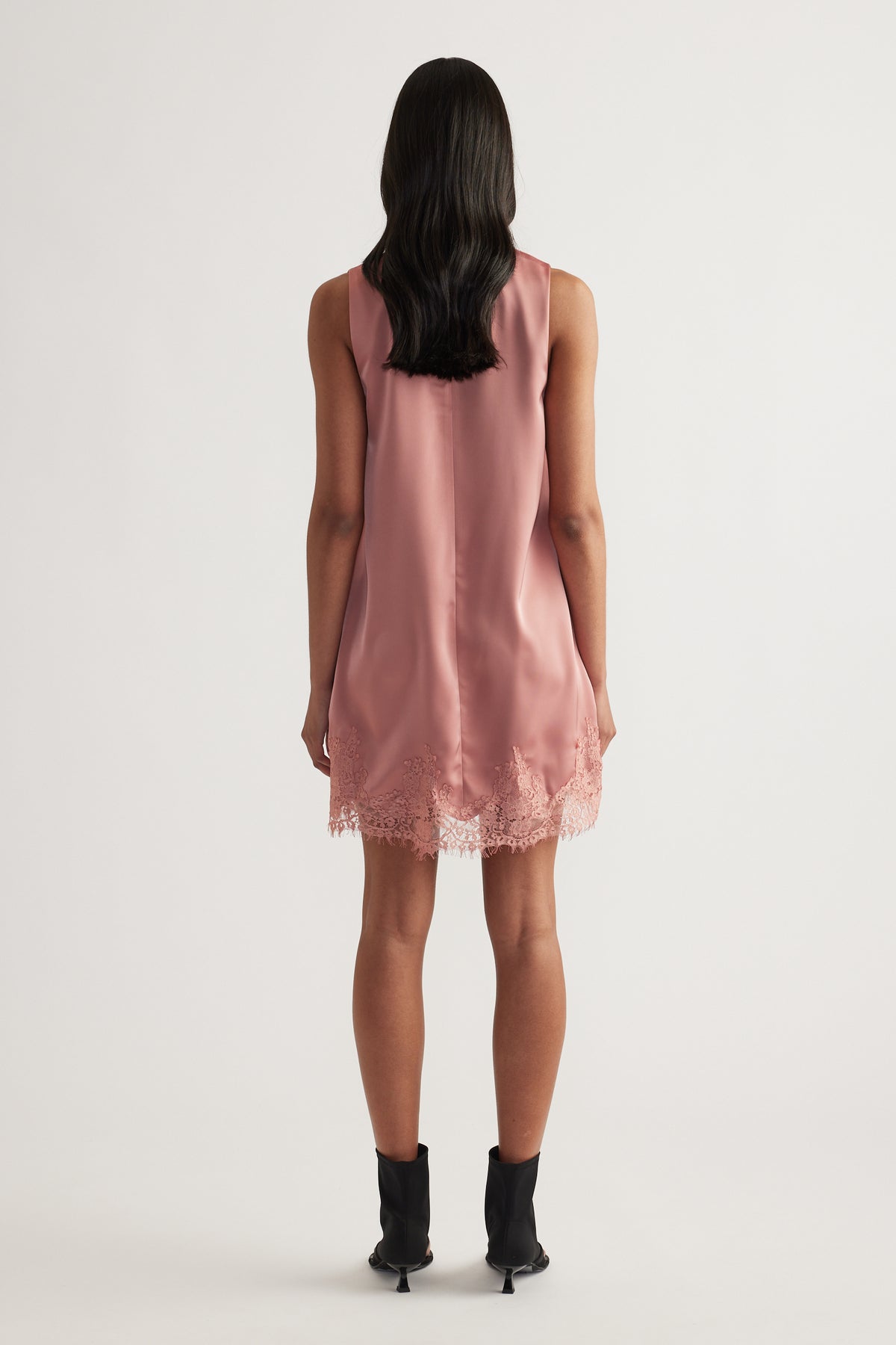 Model viewed from behind wearing a dusty rose sleeveless shift mini dress with a lace trimmed hem and black ankle boots