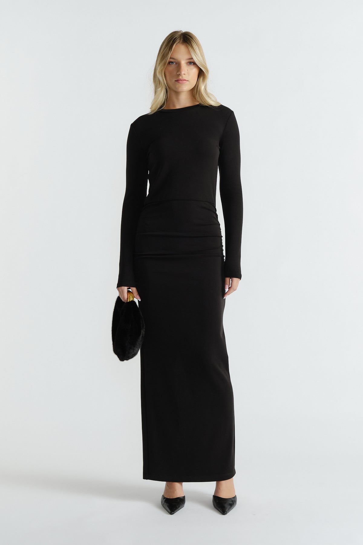 Woman wearing a sleek black long-sleeve midi dress with a fitted waist, paired with pointed black heels and a small black bag.