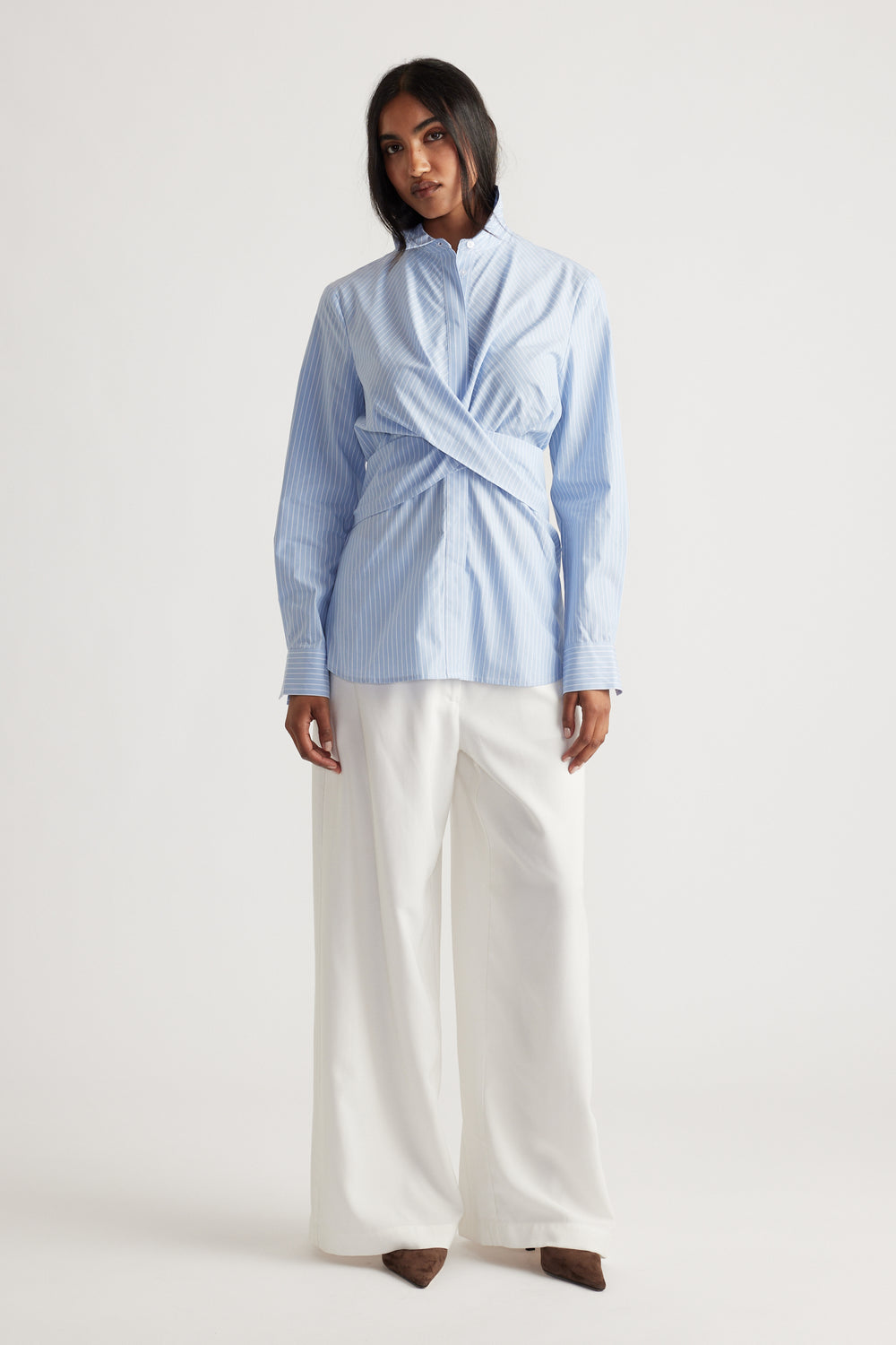 Woman wearing a light blue striped shirt with crisscross front detail paired with wide-leg white trousers and brown shoes.