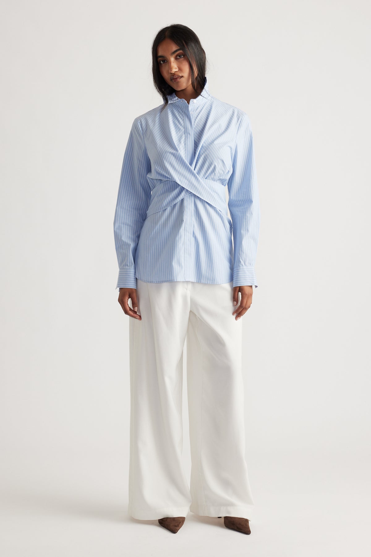 Woman wearing a light blue striped shirt with crisscross front detail paired with wide-leg white trousers and brown shoes.