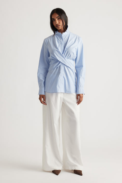 Woman wearing a light blue striped shirt with crisscross front detail paired with wide-leg white trousers and brown shoes.