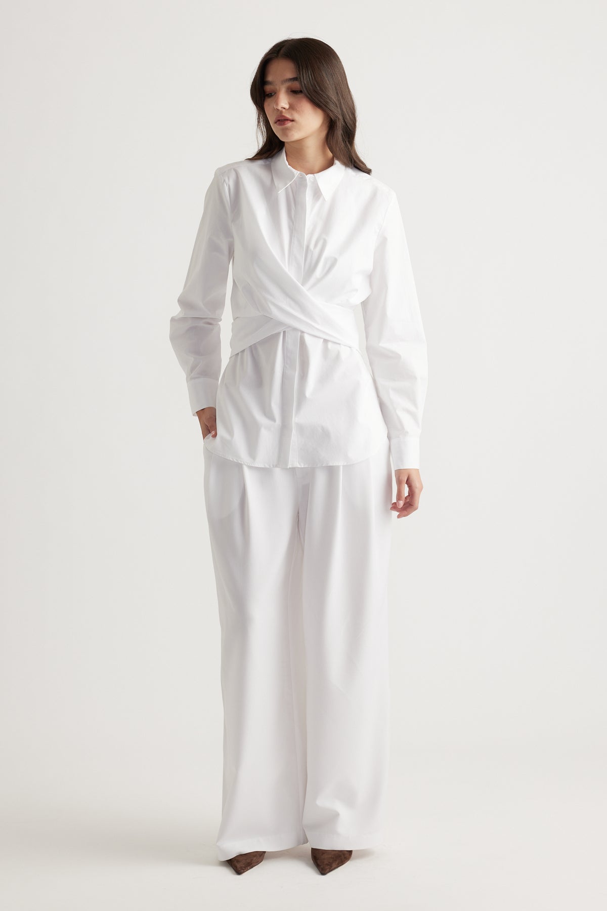 Woman wearing an elegant white long-sleeve shirt with a twist detail paired with wide-leg white trousers and brown shoes.