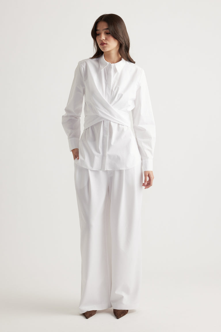 Woman wearing an elegant white long-sleeve shirt with a twist detail paired with wide-leg white trousers and brown shoes.