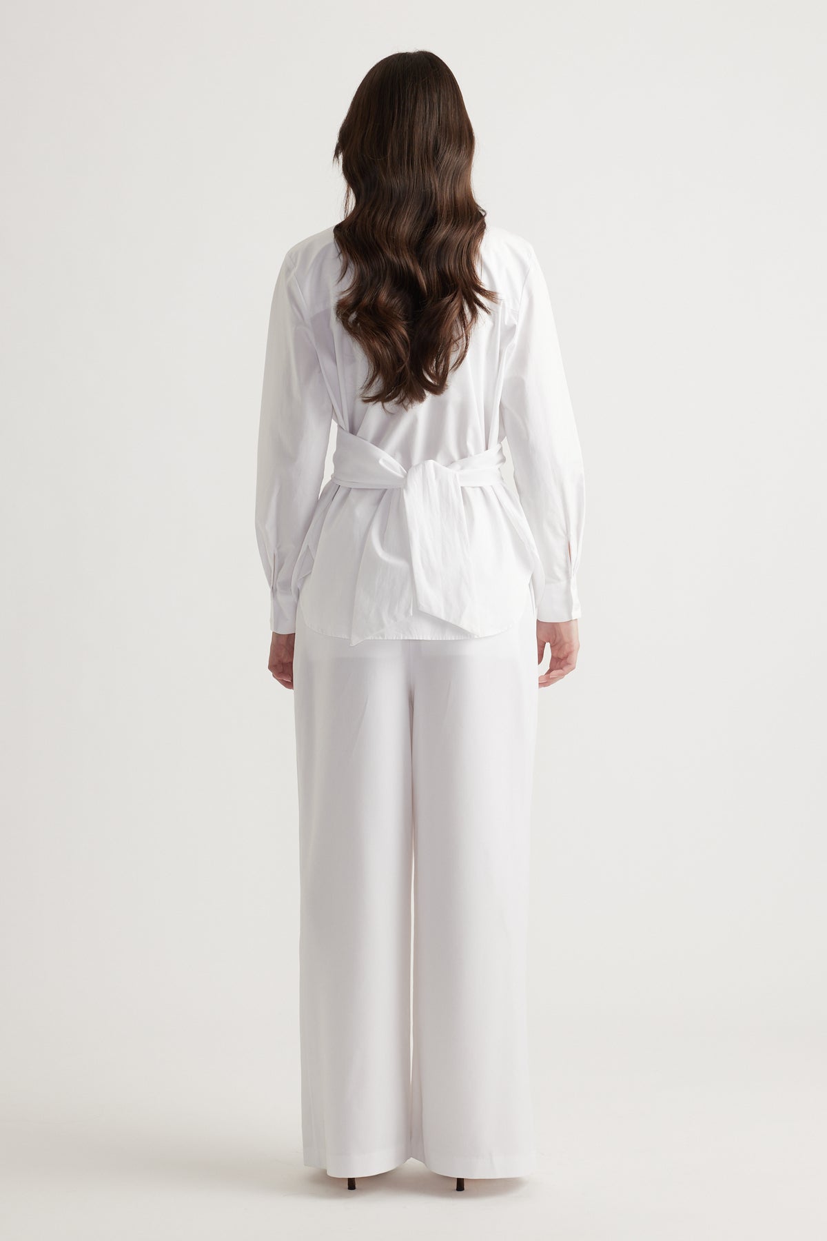 Back view of a woman wearing a white tie-back shirt paired with wide-leg white pants for a clean, elegant look.