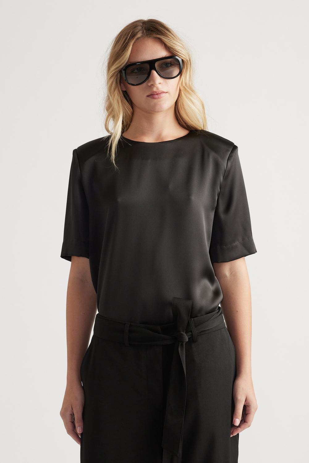 Woman wearing a black satin top with subtle shoulder pads paired with high-waisted belted trousers and oversized sunglasses