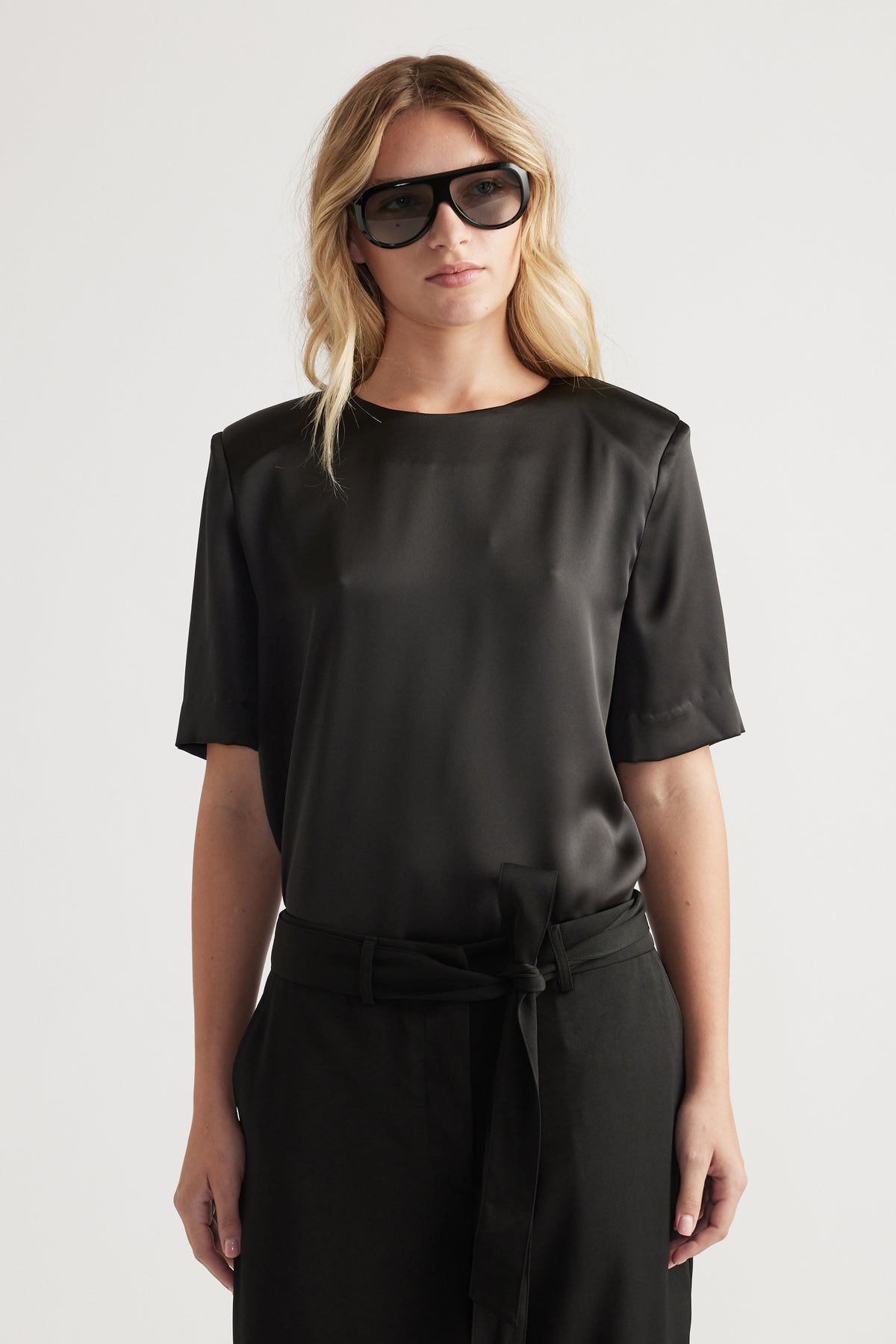 Woman wearing a black satin top with subtle shoulder pads paired with high-waisted belted trousers and oversized sunglasses