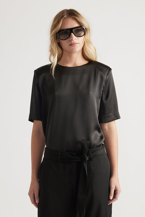 Woman wearing a black satin top with subtle shoulder pads paired with high-waisted belted trousers and oversized sunglasses