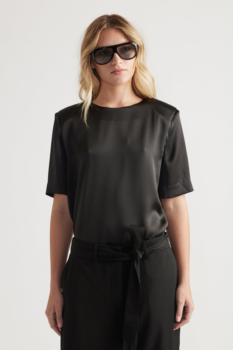 Woman wearing a black satin top with subtle shoulder pads paired with high-waisted belted trousers and oversized sunglasses