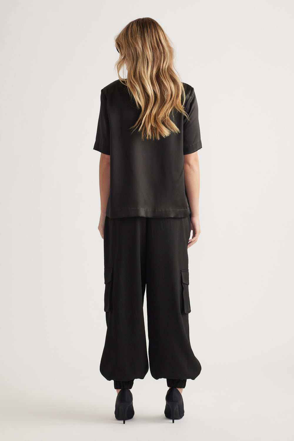 Woman seen from behind wearing a loose black satin top paired with wide-leg black cargo pants and black heels