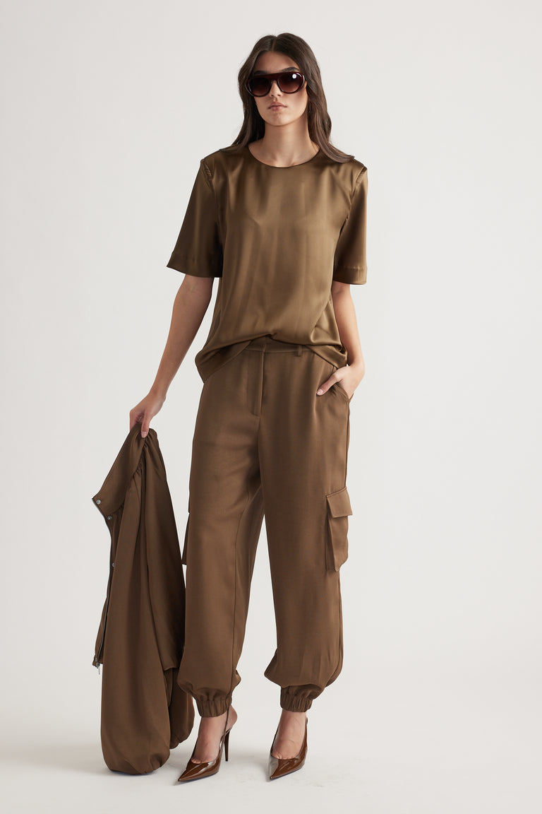 Woman wearing a bronze-toned satin top with relaxed cargo pants and pointed heels, holding a matching jacket and sunglasses.
