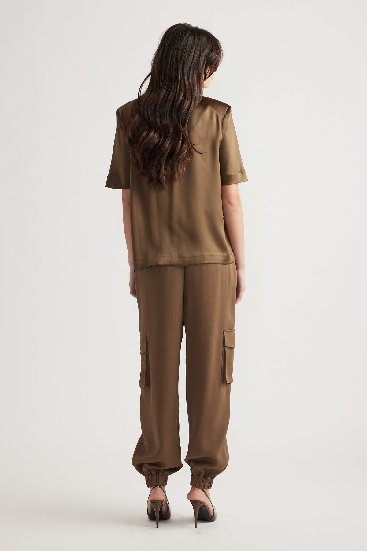 Woman wearing a bronze satin top with shoulder pads paired with relaxed cargo pants and black heeled sandals, shown from back view.