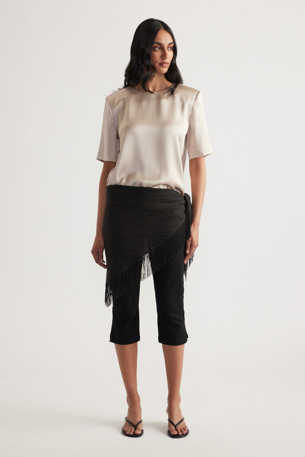 Woman wearing a satin shoulder pad top with black cropped pants and a fringed wrap skirt overlay