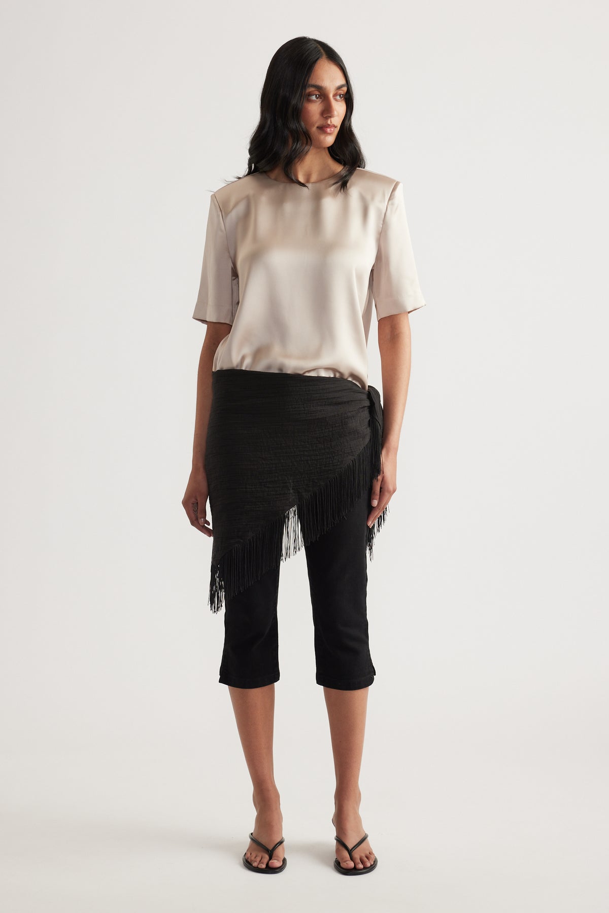 Woman wearing a satin shoulder pad top with black cropped pants and a fringed wrap skirt overlay