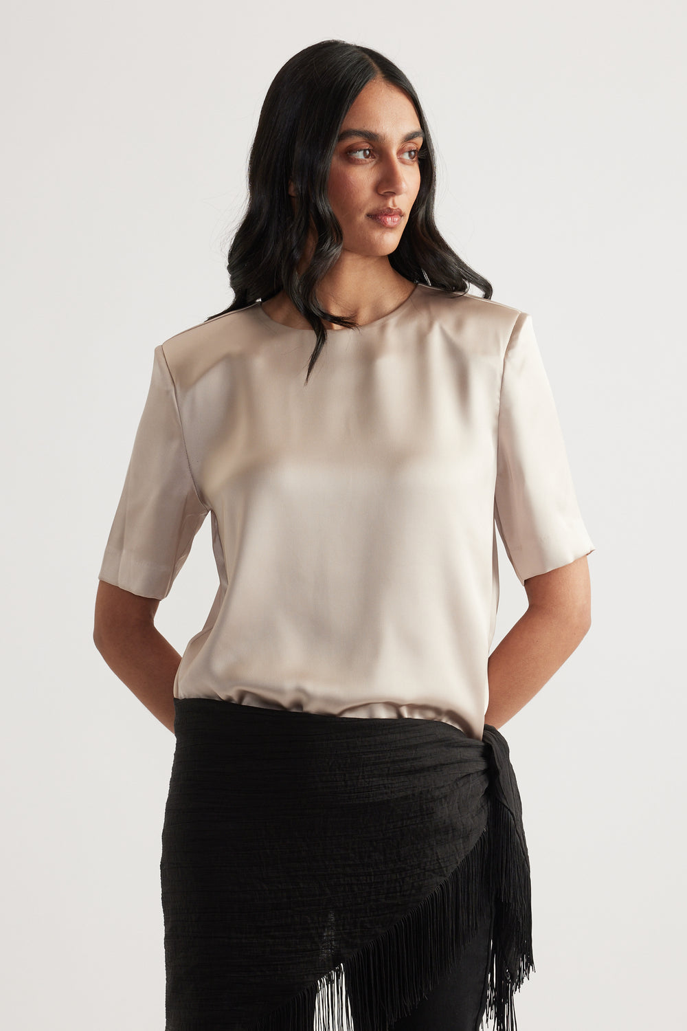 Woman wearing a taupe satin top with shoulder pads and short sleeves, styled with a black fringed wrap skirt