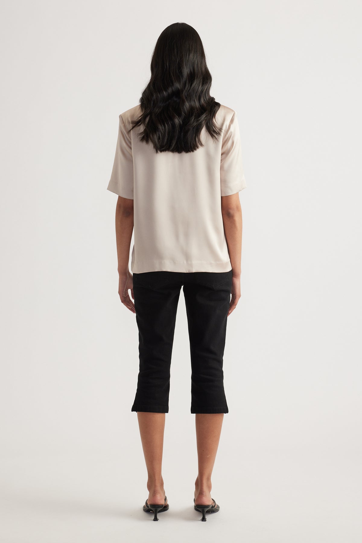 Woman viewed from behind wearing a cream satin short sleeve top with black cropped pants and heels
