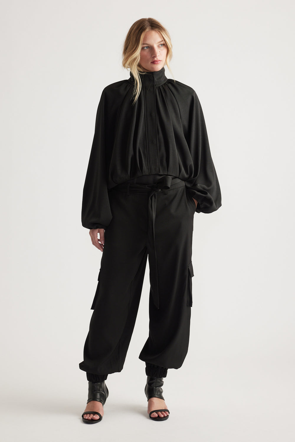 Woman wearing flowing black blouse with high neckline paired with relaxed fit black satin cargo pants and heels