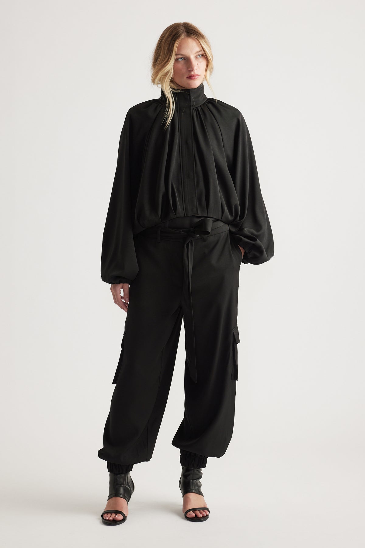 Woman wearing flowing black blouse with high neckline paired with relaxed fit black satin cargo pants and heels